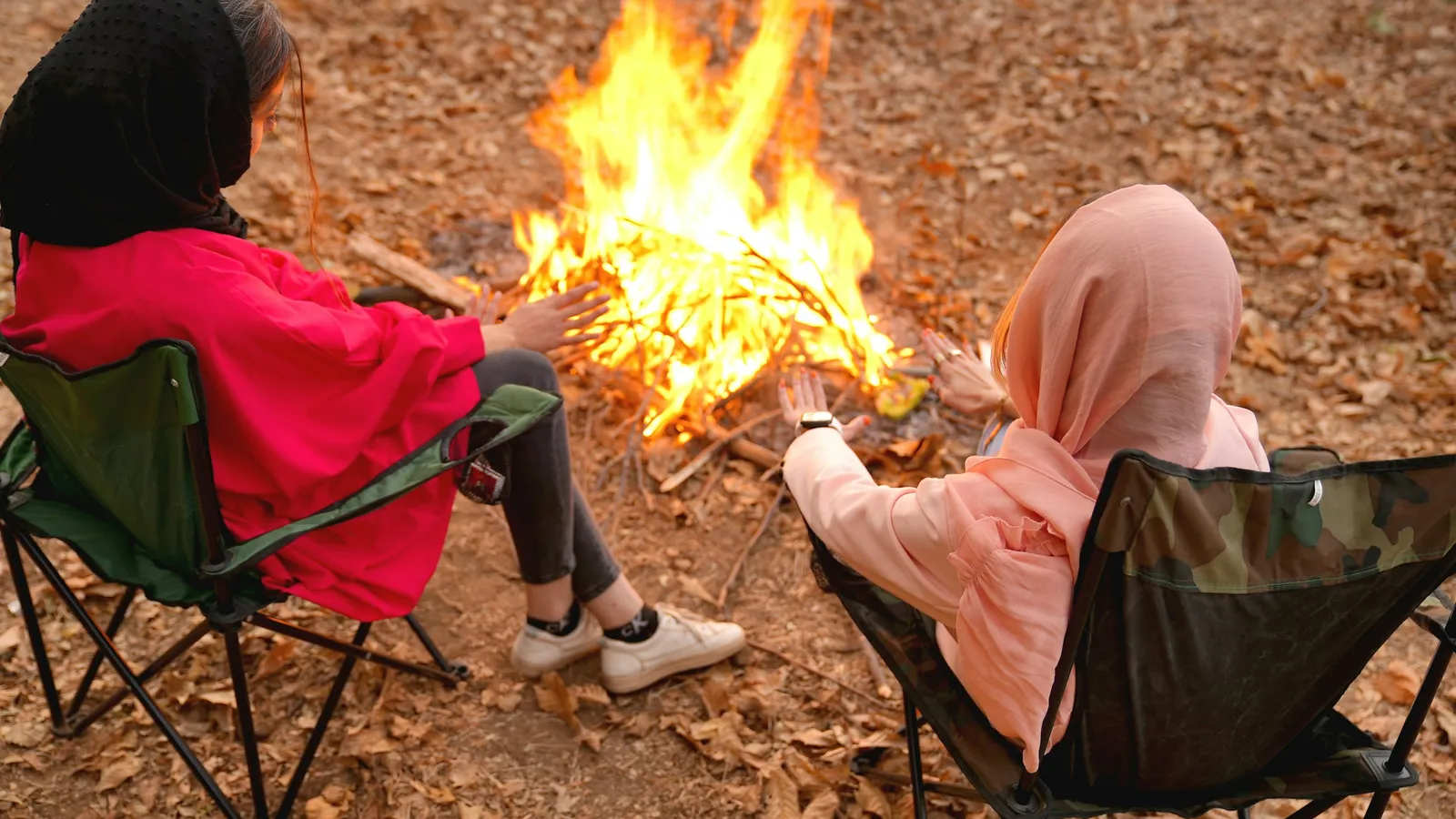 Two people in folding chairs around a campfire in autumn woods